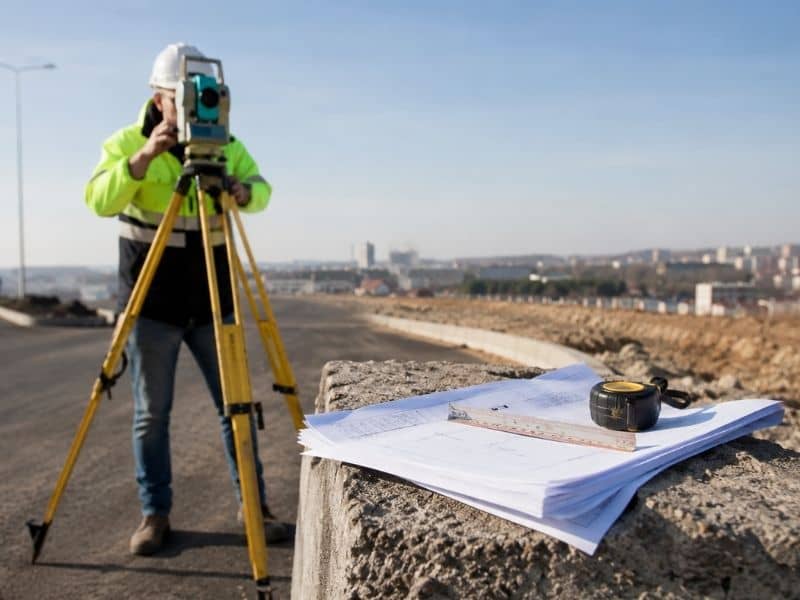 A licensed surveyor in a high-visibility jacket using a total station on a lot with blueprints in the foreground illustrating property survey cost considerations